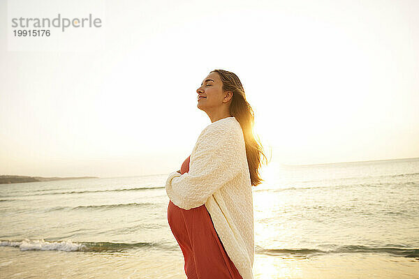 Glückliche schwangere Frau steht mit geschlossenen Augen am Strand