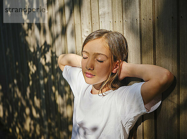 Mädchen mit geschlossenen Augen lehnt an einer Holzwand und genießt das Sonnenlicht