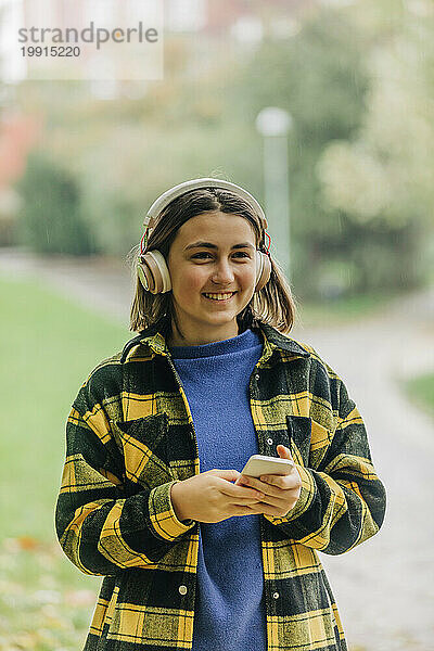 Lächelndes Teenager-Mädchen hält Smartphone in der Hand und hört Musik über kabellose Kopfhörer im Park