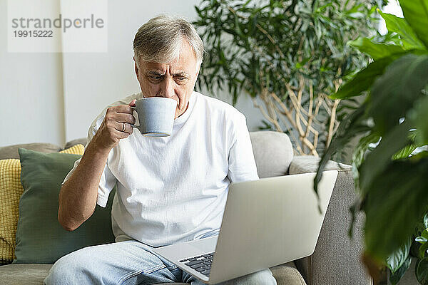 Älterer Mann trinkt Kaffee und benutzt Laptop zu Hause