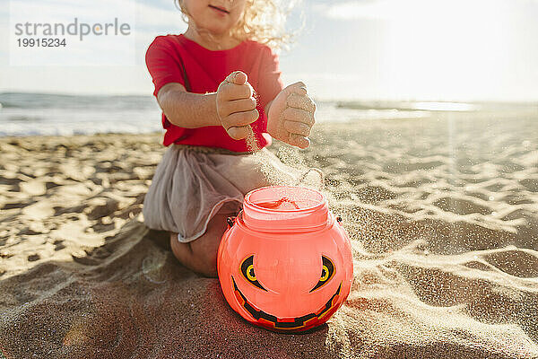 Mädchen füllt an einem sonnigen Tag am Strand Sand in Halloween-Kürbisspielzeug