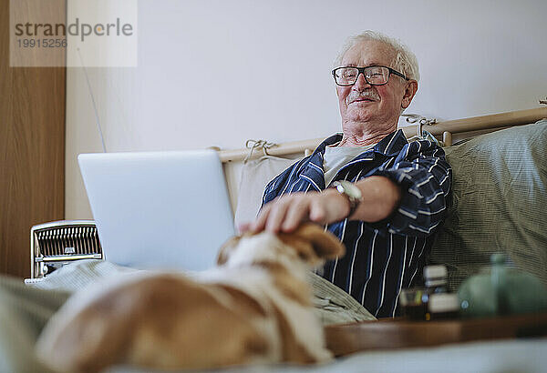 Glücklicher älterer Mann sitzt mit Laptop und streichelt Hund zu Hause im Bett