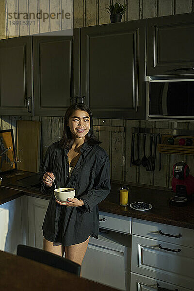 Glückliche junge Frau hält eine Schüssel Frühstück in der Hand und steht zu Hause in der Küche