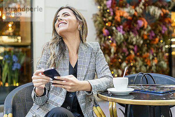 Glückliche Geschäftsfrau mit Smartphone sitzt im Straßencafé