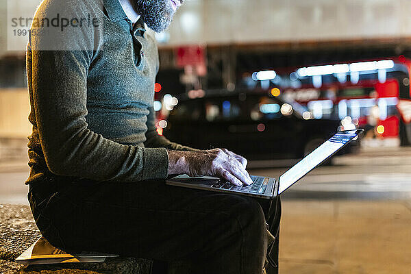 Freelancer using laptop sitting on bench in city at night