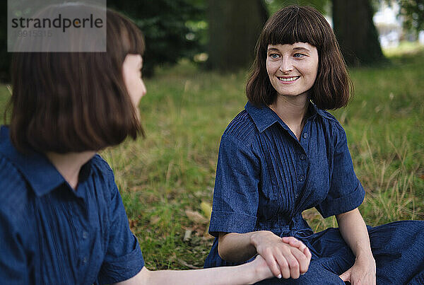 Glückliche Zwillingsschwestern halten Händchen und sitzen auf Gras