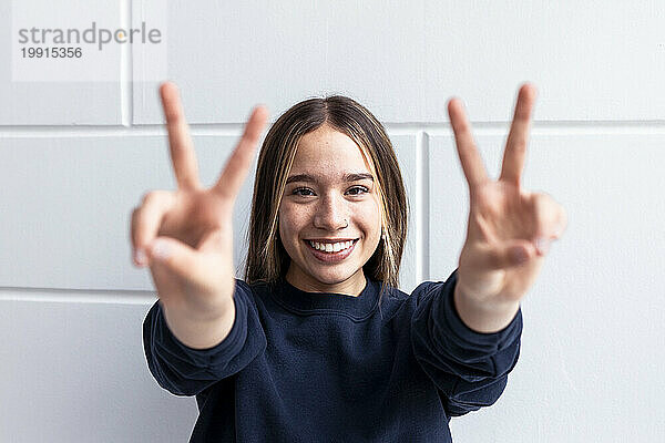 Smiling woman gesturing peace sign in front of wall at home