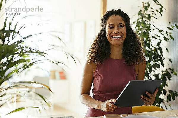 Glückliche Geschäftsfrau hält Tablet-PC in der Nähe von Pflanzen im Büro