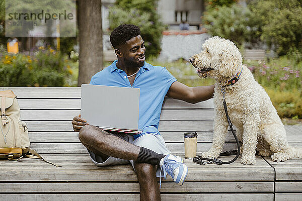 Lächelnder junger Mann mit Laptop streichelt Hund  während er auf Bank im Park sitzt