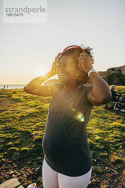 Glückliche schwangere Frau  die bei Sonnenuntergang kabellose Kopfhörer trägt