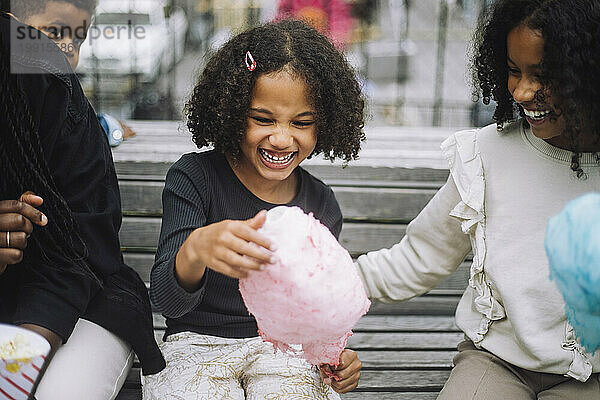 Fröhliches Mädchen  das Spaß mit Geschwistern hat  während es Zuckerwatte im Vergnügungspark hält