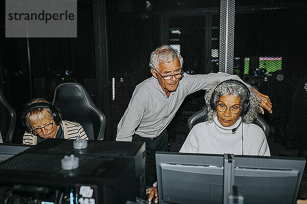Elderly woman playing video game on computer by male and female friends at gaming lounge