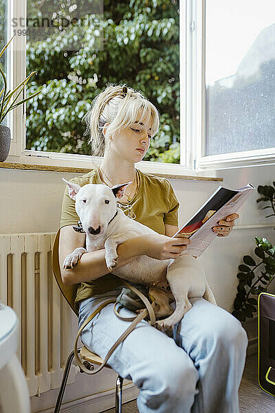 Blonde Frau mit Bullterrier-Hund liest Zeitschrift auf Stuhl im Wartezimmer einer Tierklinik
