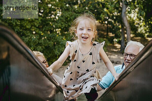 Porträt eines lächelnden Mädchens  das auf einer Rutsche klettert  während es mit Großeltern im Park spielt