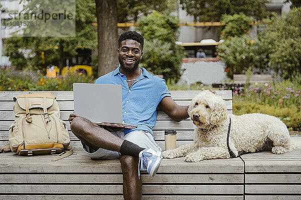 Porträt eines lächelnden Mannes mit Laptop  der neben einem Labradoodle-Hund auf einer Bank im Park sitzt