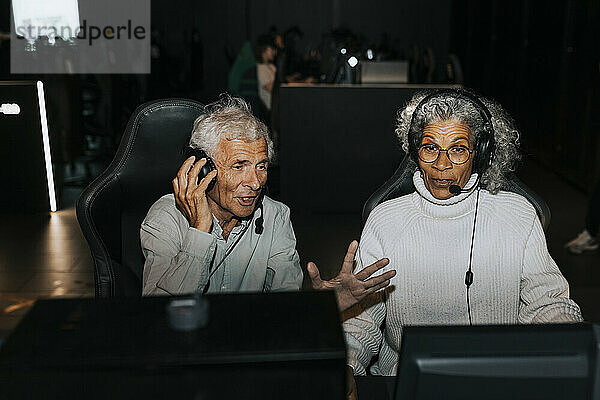 Senior man wearing headset sitting by female friend playing video game on computer at gaming lounge