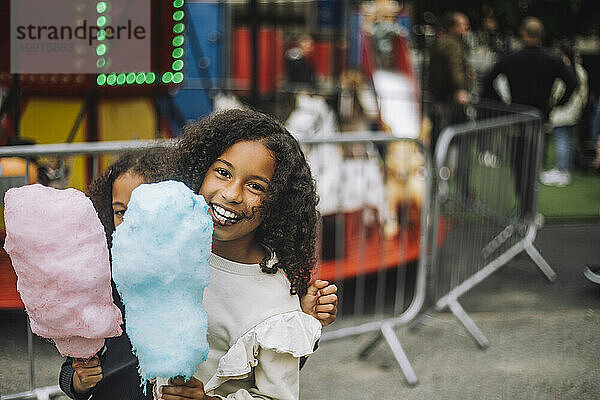 Porträt eines glücklichen Mädchens  das mit seiner Schwester Zuckerwatte im Vergnügungspark hält