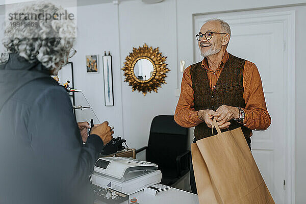 Lächelnder älterer männlicher Unternehmer  der einer Kundin beim Einkaufen im Geschäft eine Papiertüte schenkt