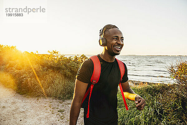 Glücklicher junger Mann mit Rucksack und kabellosen Kopfhörern  der auf dem Fußweg läuft