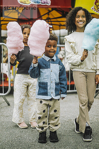 Porträt glücklicher Geschwister  die Zuckerwatte halten  während sie im Vergnügungspark stehen