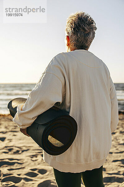 Rückansicht einer älteren Frau  die am Strand steht und eine Trainingsmatte in der Hand hält und aufs Meer blickt