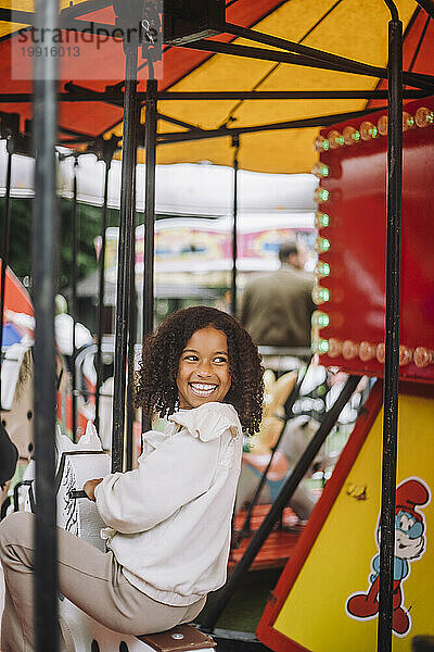 Glückliches Mädchen  das über die Schulter schaut  während es Spaß auf dem Karussell im Vergnügungspark hat