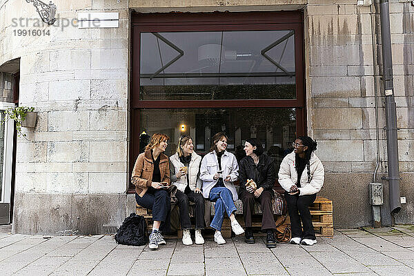 Junge  glückliche Freundinnen reden miteinander  während sie auf Paletten am Bürgersteig in der Stadt sitzen