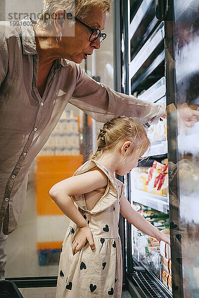 Seitenansicht eines Mädchens mit der Hand auf der Hüfte beim Einkaufen im Kühlregal in der Nähe der Großmutter im Geschäft