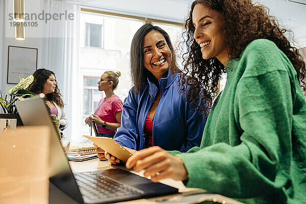 Glückliche Geschäftsfrau mit lächelnder Kollegin  die im Büro auf den Laptop schaut