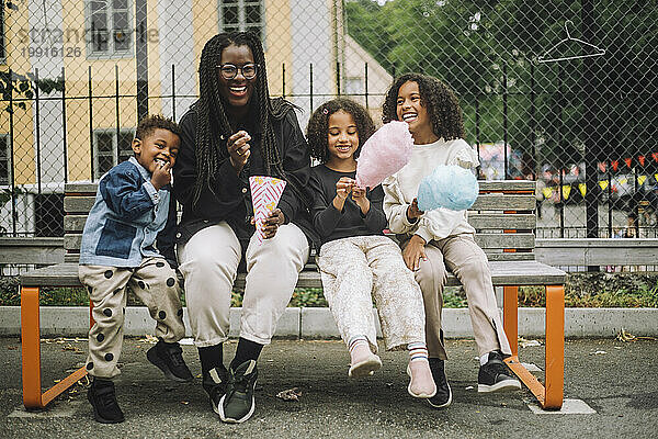 Porträt glücklicher Kinder  die Zuckerwatte essen  während sie auf einer Bank im Vergnügungspark sitzen