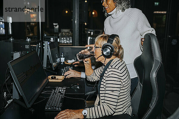 Ältere Frau trinkt Wasser  während sie von einer Freundin im Gaming-Center ein Computerspiel spielt