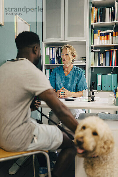 Junger Mann mit Hund spricht mit einer Tierärztin am Schreibtisch in der Klinik