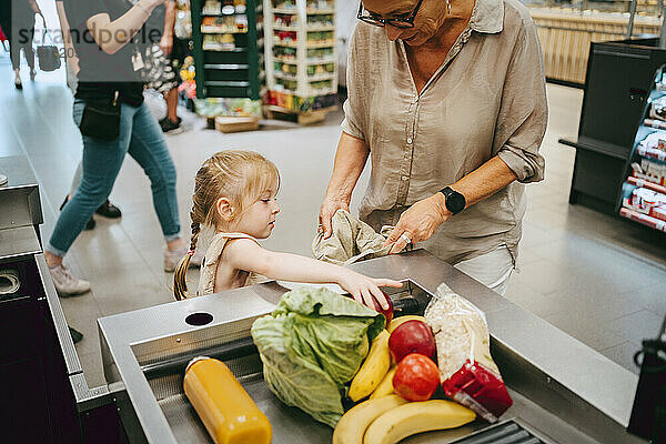 Mädchen hilft Großmutter beim Befüllen von Einkaufstüten an der Kasse im Supermarkt