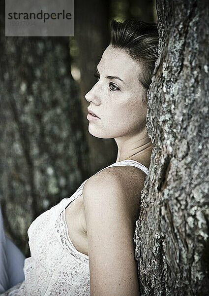 Eine junge Frau in einem weißen Tanktop lehnt mit dem Rücken an einem Baum.
