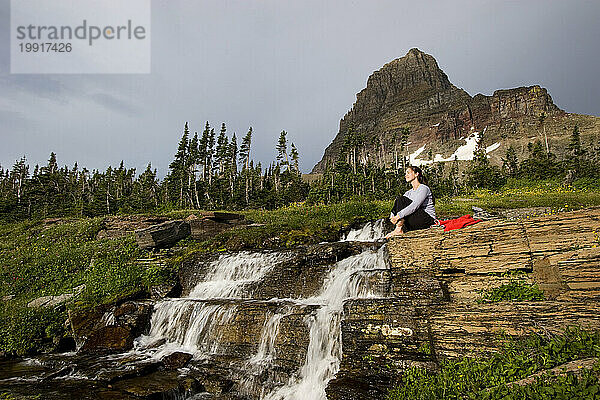 Eine erwachsene Frau sitzt nachdenklich mit Clement Mountai auf dem Felsvorsprung eines kleinen Wasserfalls auf dem Highline-Trail in der Nähe des Logan Pass