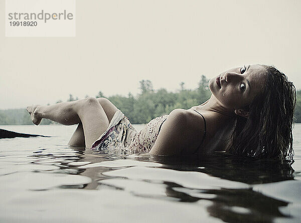 Eine junge Frau in einem weißen Kleid entspannt sich halb untergetaucht in einem See in Maine.
