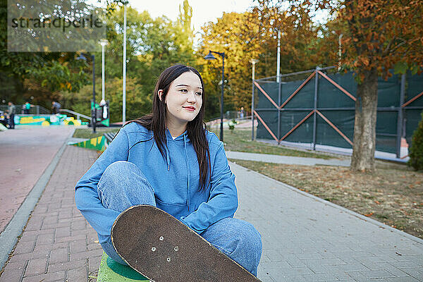 Lächelndes junges Mädchen mit einem Skateboard  das im Park sitzt