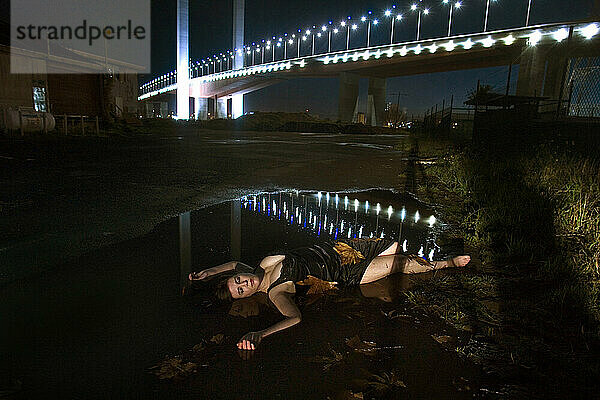 Eine Frau in einem Kleid liegt nachts in einer Pfütze unter einer Brücke in Australien.