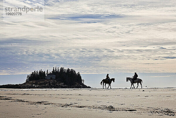 Zwei Personen reiten auf Pferden einen Sandstrand in Maine entlang.