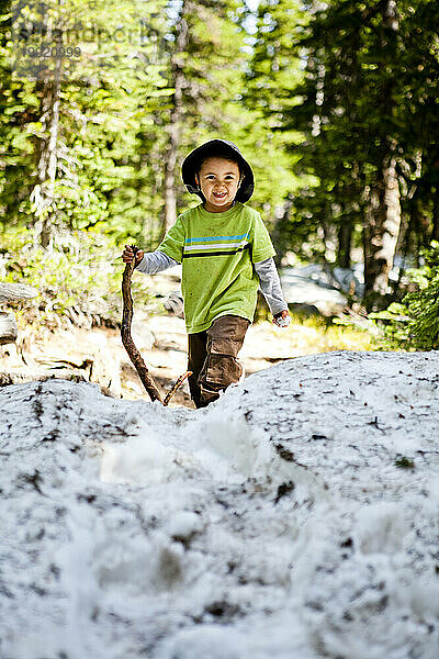 Ein 3-jähriger Junge wandert mit einem Stock als Gleichgewicht über sommerliche Schneeverwehungen.
