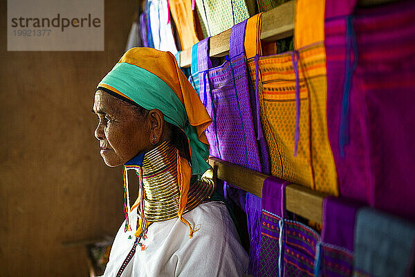 Portrait of senior woman wearing traditional neck rings standing beside colorful clothes hanging on wooden beams  Shan State  Myanmar