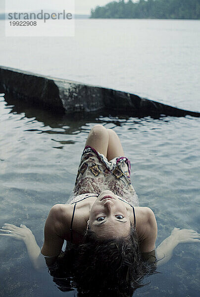 Eine junge Frau in einem weißen gemusterten Kleid entspannt sich im klaren Wasser eines Sees in Maine.