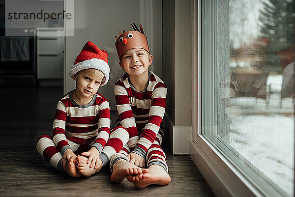 Zwei kleine Jungen sitzen im Weihnachtspyjama neben dem verschneiten Fenster