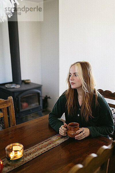 Frau sitzt mit Kaffeetasse am Küchentisch in gemütlicher Hütte