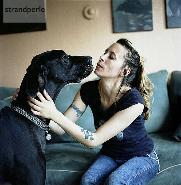 Young woman kisses her dog's nose while sitting on the couch at home (great dane).