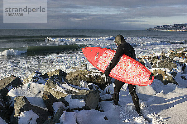 Mit seinem Surfbrett macht sich ein junger Mann auf den Weg durch den Schnee in Richtung kalter Gewässer in Homer  Alaska.