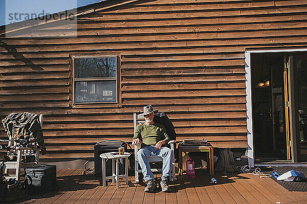 Mann mit Cowboyhut sitzt im Schaukelstuhl auf der Veranda einer Holzhütte