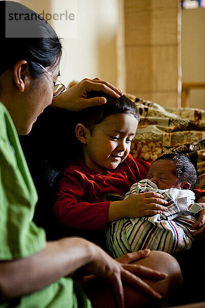 Ein kleiner Junge hält seinen neugeborenen kleinen Bruder  während seine Mutter zuschaut.