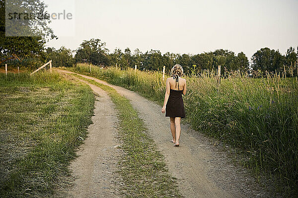 Ein Mädchen in einem Kleid geht eine unbefestigte Straße entlang und durch ein Feld im ländlichen Maine.