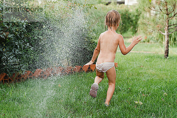 Glückliches 3-jähriges Mädchen wird nur in Höschen unter einer Sprinkleranlage nass